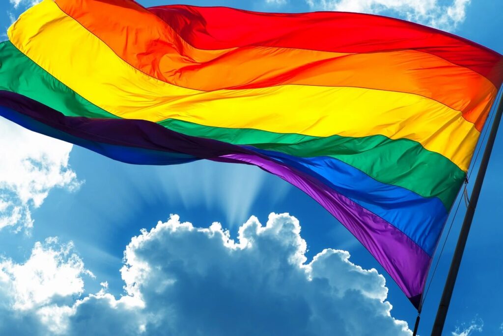 Bright rainbow flag waving against a blue sky with clouds.